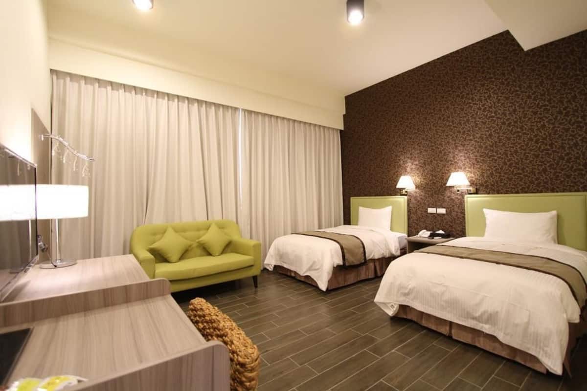A hotel room with two beds, a green sofa, a desk with a lamp, and a large window with curtains.