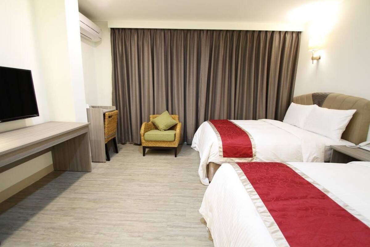A hotel room with two beds, a television, a desk, a chair, and a large window with curtains.