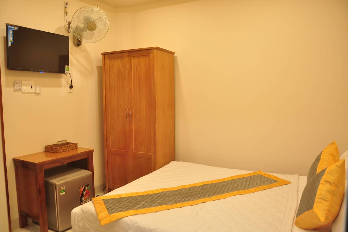 Un dormitorio con cama, un armario de madera, un refrigerador pequeño, un televisor montado en la pared y un ventilador de pared.
