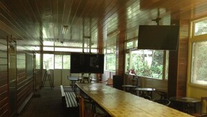 A long dining table with chairs, a television mounted on the wall, and large windows with a view of greenery outside.