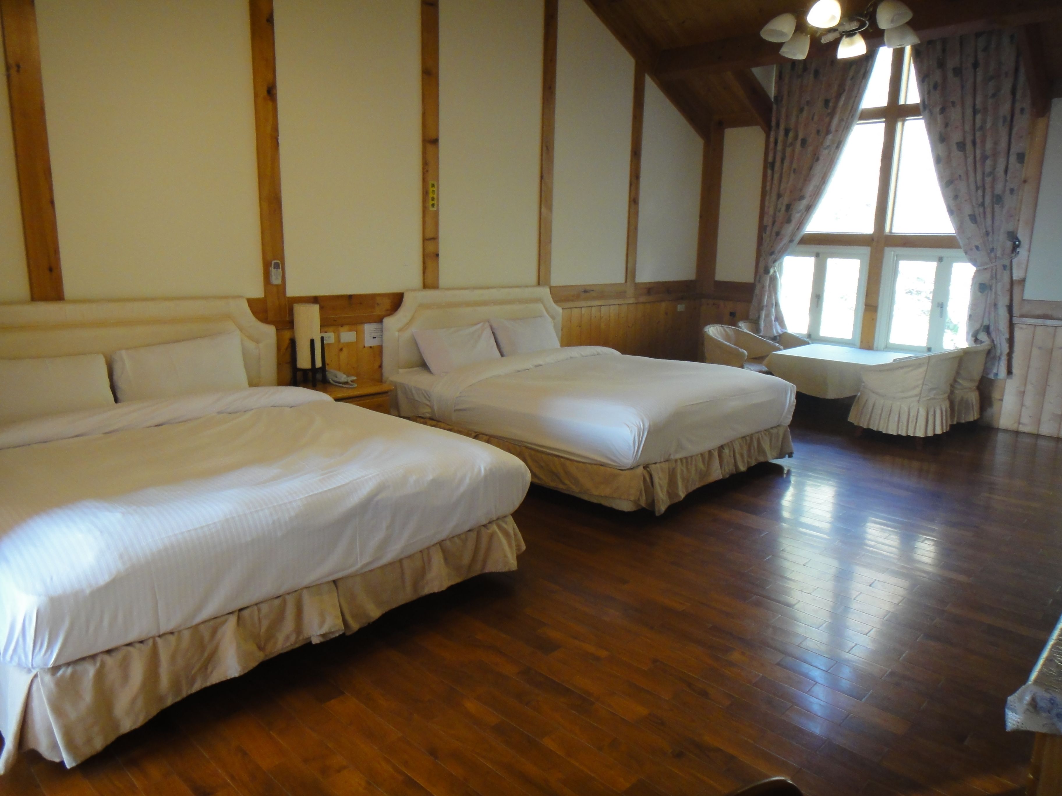 A room with two beds, wooden floors, a window with curtains, and a small table with chairs.