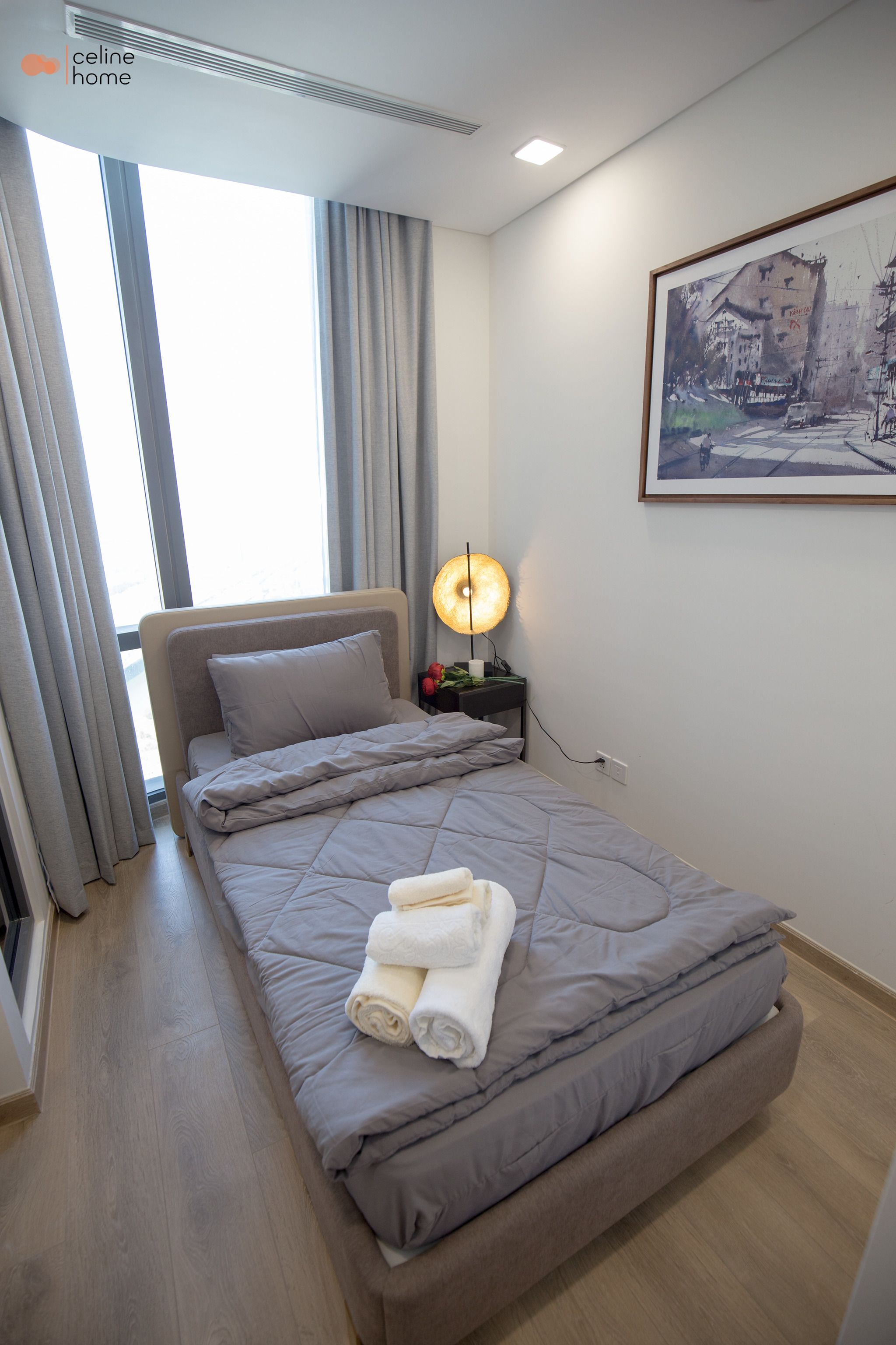 A modern hotel room with a large bed, a bedside lamp, and a framed cityscape painting on the wall.