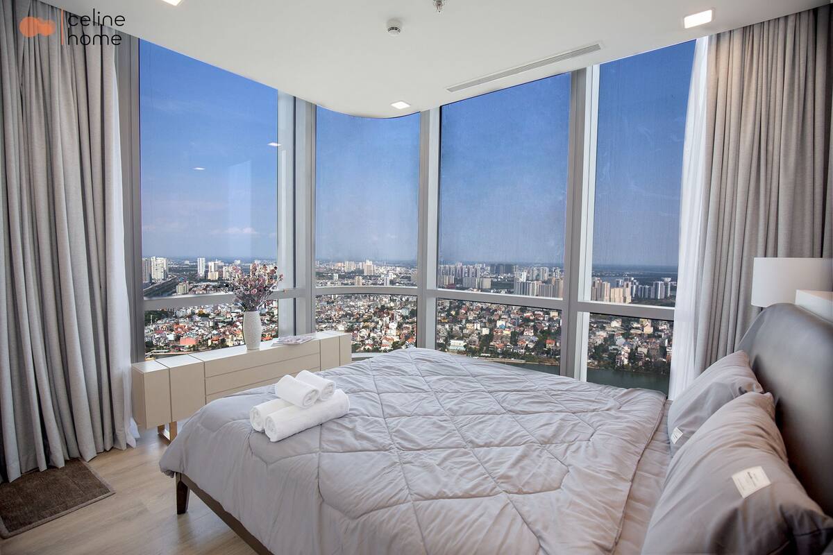 Quarto moderno com uma cama grande, uma mesa de cabeceira e vista para a cidade através de grandes janelas.