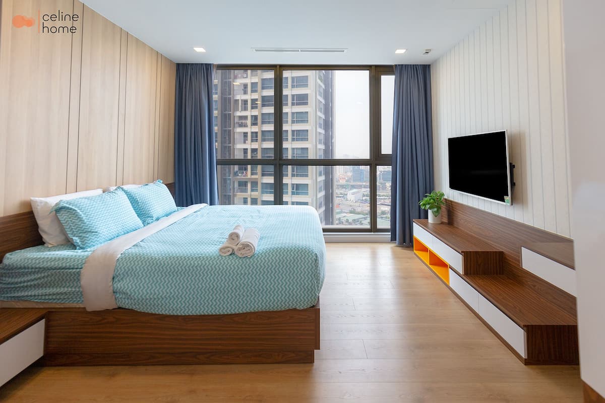 Quarto moderno com uma janela grande, cama com roupa de cama azul, televisão na parede e um banco de madeira com uma planta.