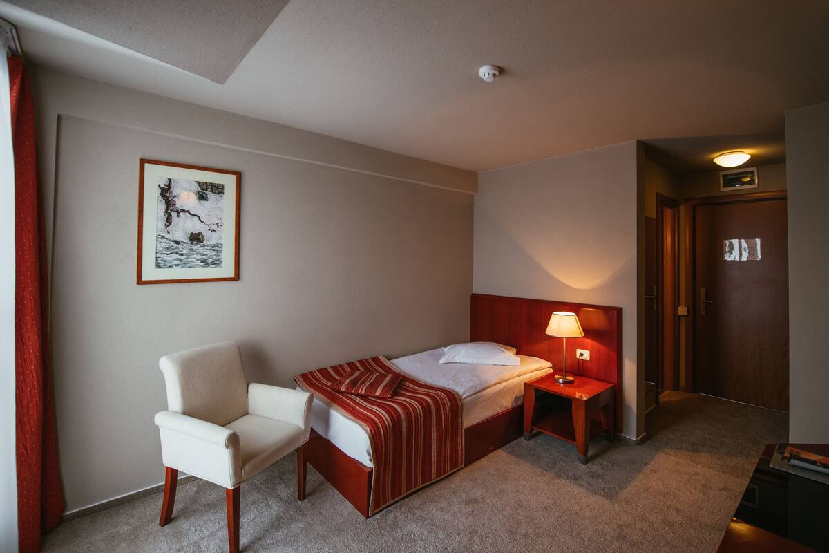 A hotel room with a bed, a chair, a bedside table with a lamp, a framed picture on the wall, and a door.