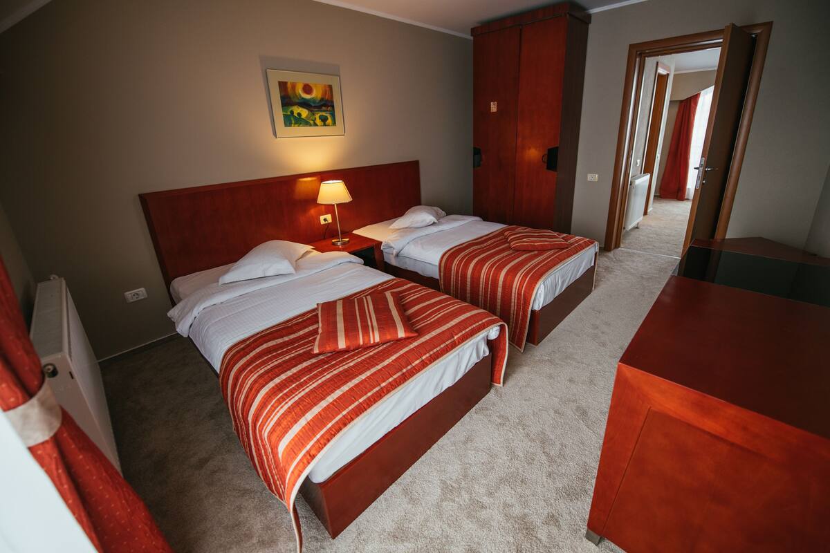 A hotel room with two beds, a wooden headboard, a nightstand with a lamp, a painting on the wall, and a wardrobe.