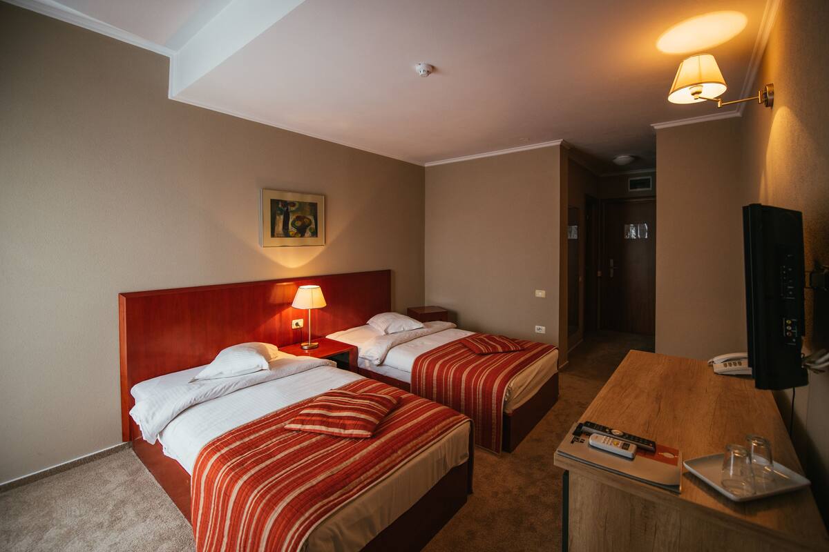 A hotel room with two beds, a wooden desk, a lamp, a painting on the wall, and a television.