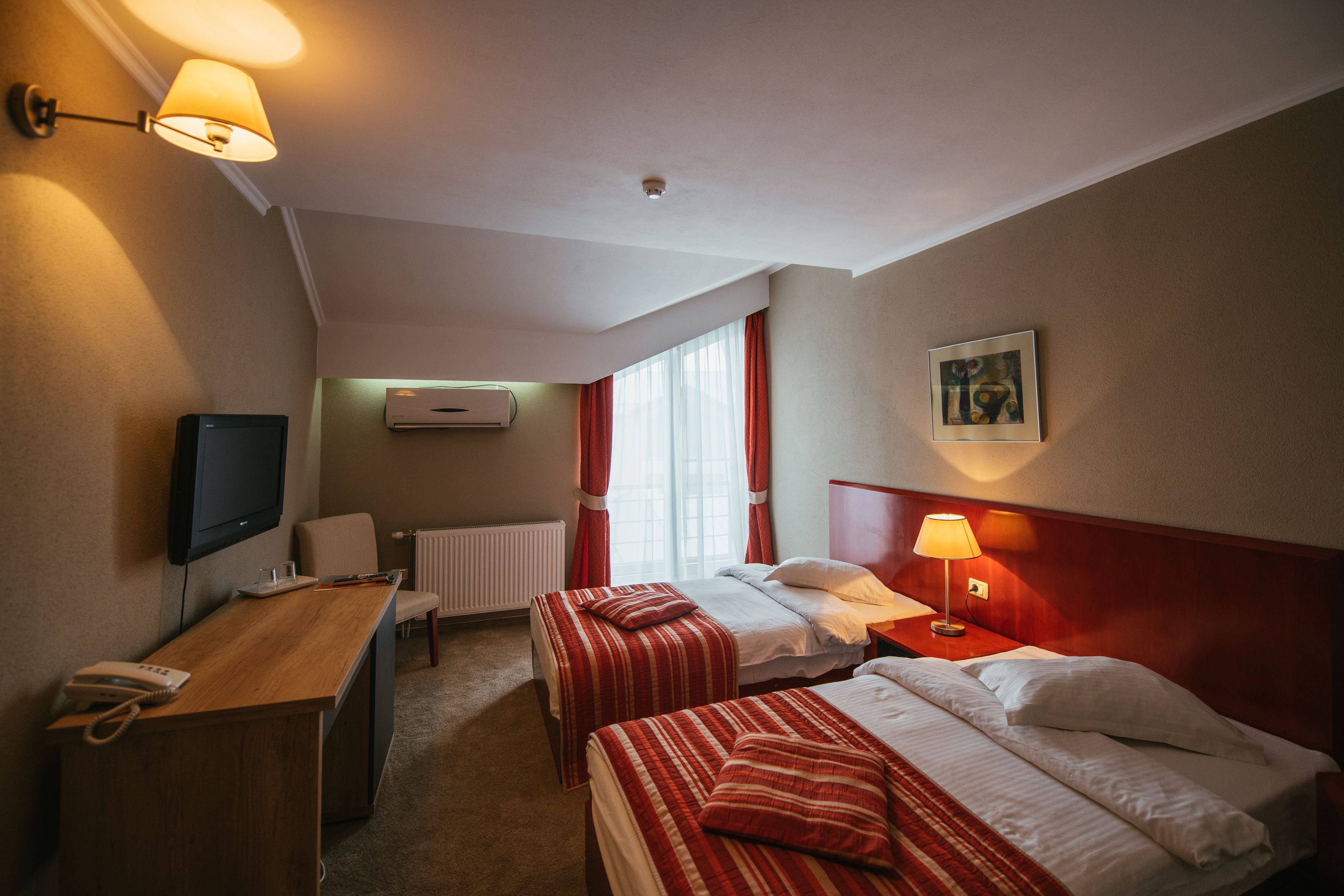 Een hotelkamer met twee bedden, een bureau met telefoon, een televisie en een raam met gordijnen.