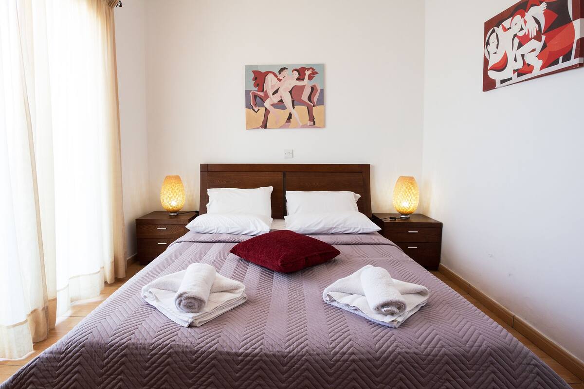 Un lit bien fait, recouvert d’une courtepointe violette, avec deux oreillers blancs et un coussin décoratif rouge. On y trouve deux lampes de chevet, une table de chevet en bois et deux tableaux accrochés au mur.