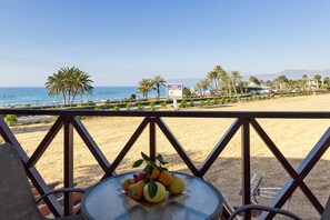 A table with a fruit bowl on a balcony overlooking a beach with palm trees and a clear sky.