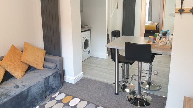 Een moderne woonkamer met een grijze bank, een salontafel en een eethoek met een tafel en stoelen. Achtergrond: een wasmachine.