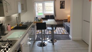 Een compacte keuken met een gasfornuis, spoelbak en moderne apparaten. Er is een eethoek met een tafel en stoelen, en een klein zithoekje met een bank.
