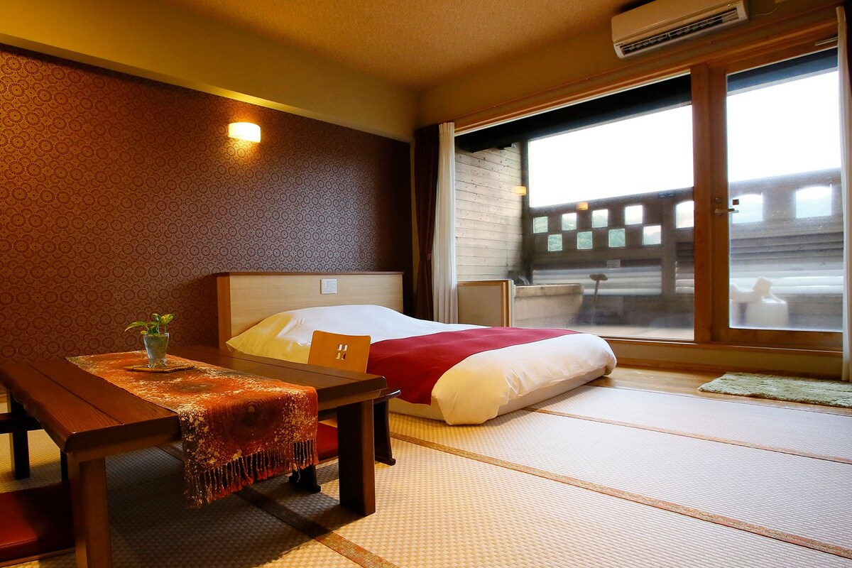 Une chambre d’hôtel avec un sol recouvert de tatamis, un lit avec une couvre-lit rouge et blanc, une table en bois ornée d’une petite plante, et une fenêtre donnant sur un immeuble.