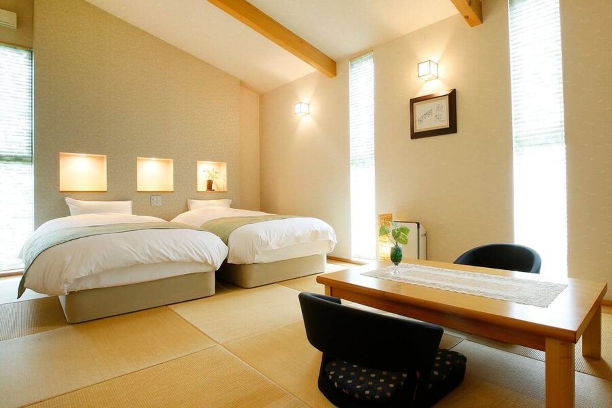 A hotel room with two beds, a dining table, tatami flooring, and wall-mounted lights.