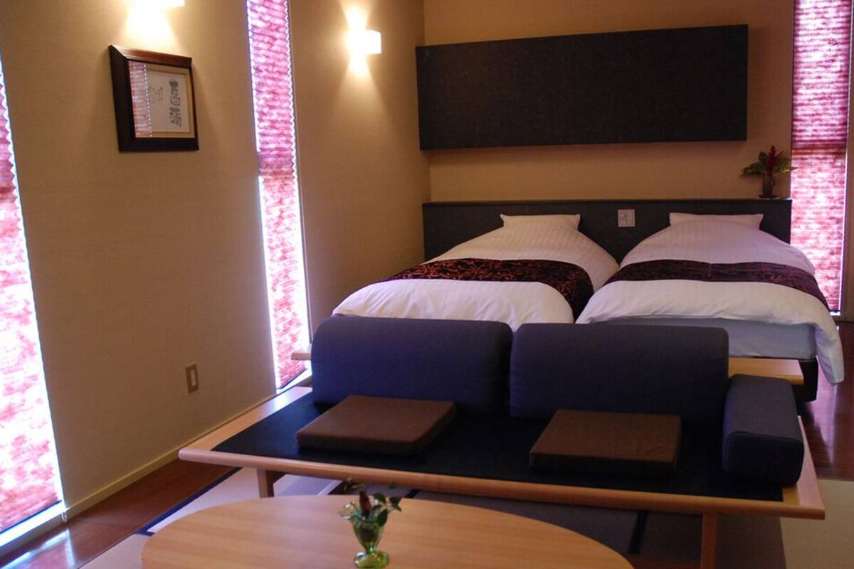 A hotel room with a bed, a sofa, a small table, and a window with blinds.