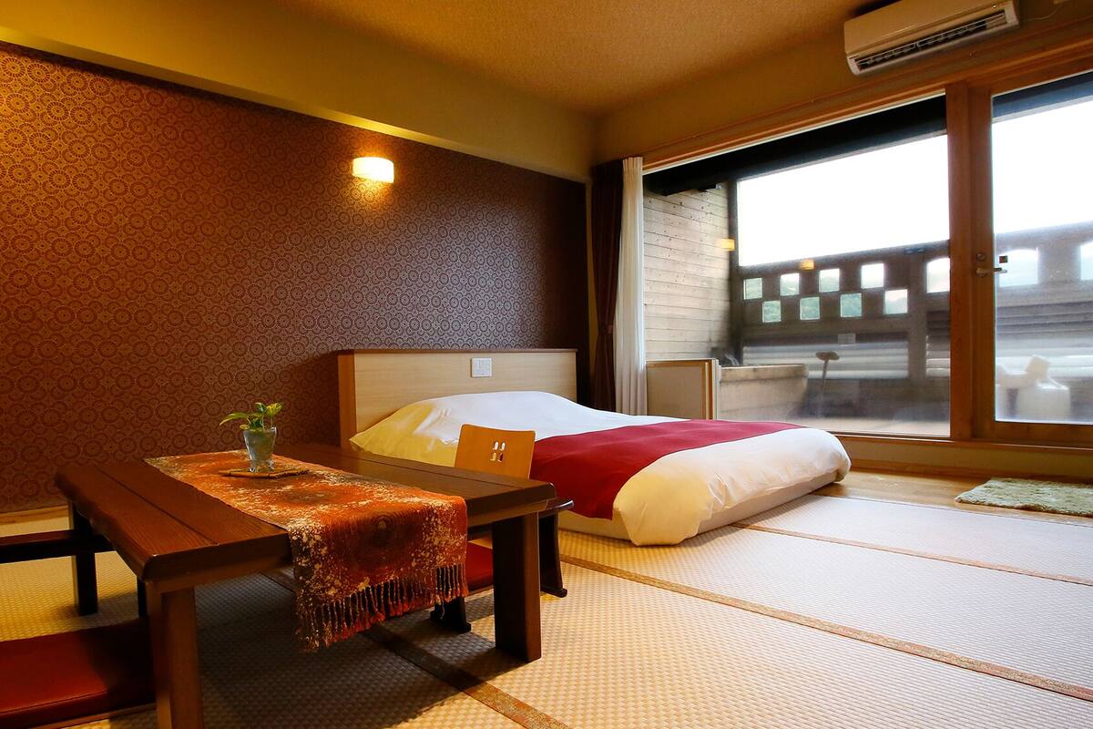 A hotel room with tatami flooring, a bed with a red and white bedspread, a wooden table with a small plant, and a window with a view of a building.