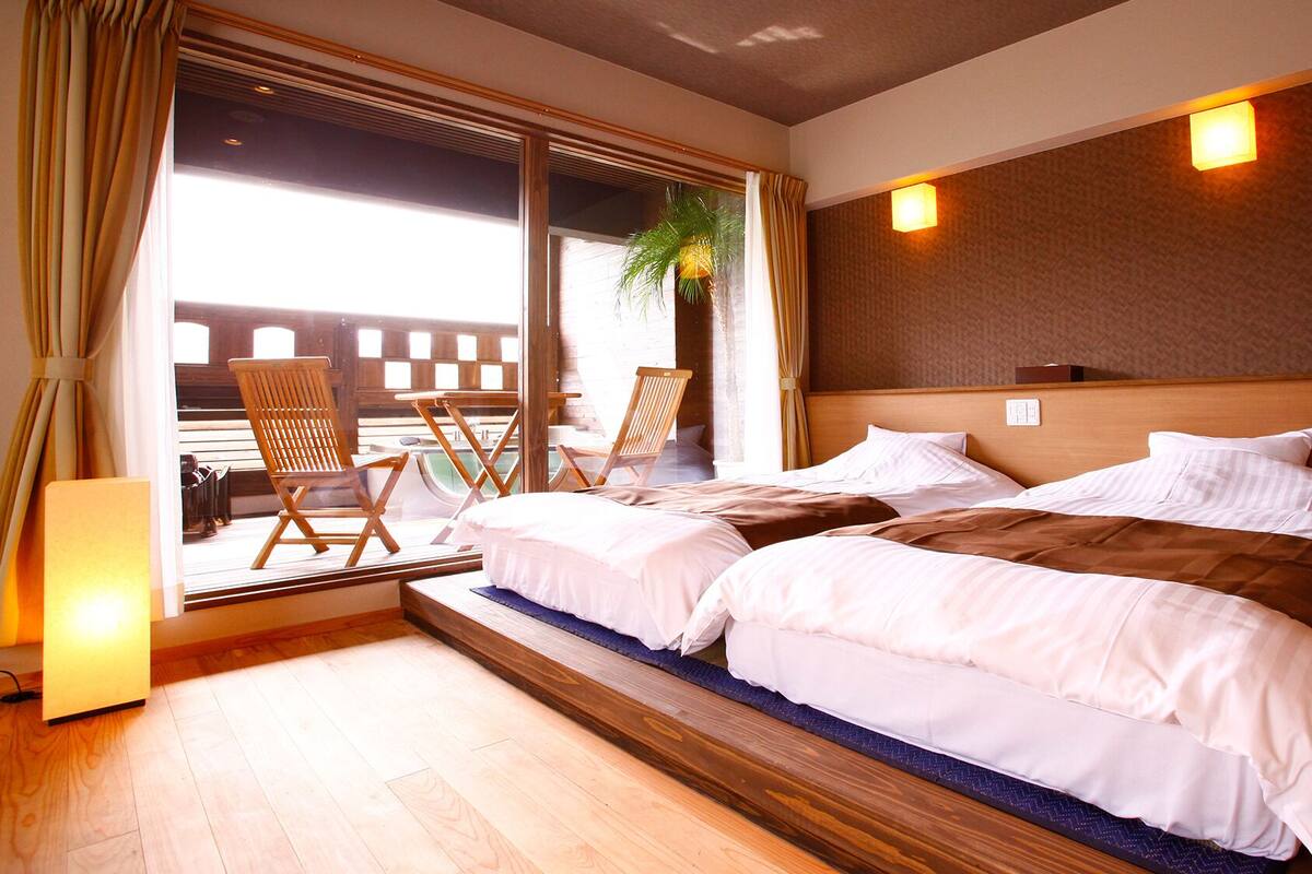 A hotel room with a large bed, two chairs, a small table, and a view of a building through a window.