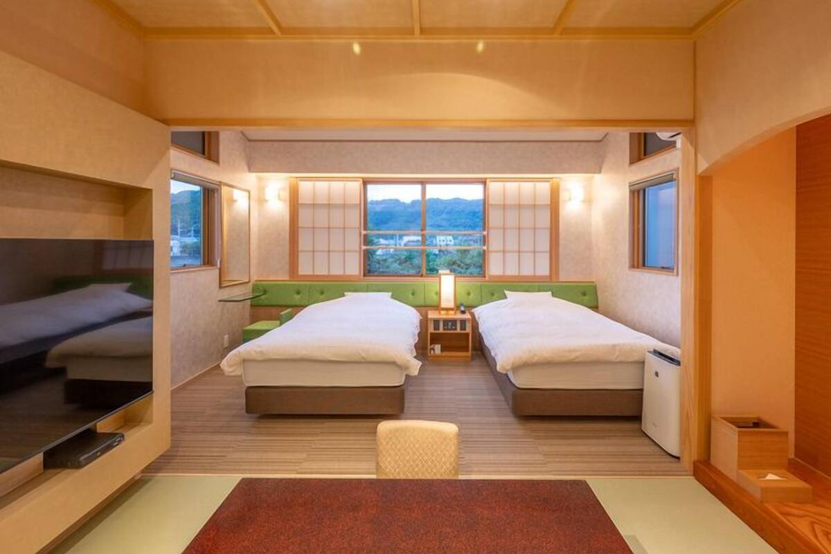 A compact hotel room with two beds, a small table, and a view of the outdoors.