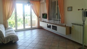 Quarto com sofá, televisão sobre suporte, radiador e janela com cortinas.