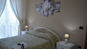Cama bem arrumada com colcha floral, duas luminárias de cabeceira e uma janela com cortinas leves.