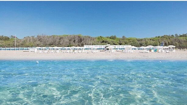 Um hotel à beira-mar com águas azul-claras, uma praia arenosa e uma fileira de edifícios brancos com guarda-sóis.
