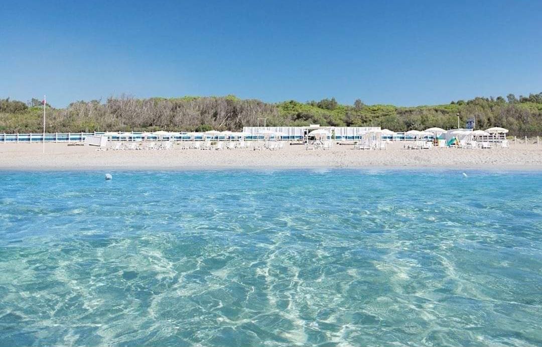 Ein Hotel am Strand mit klarem, blauem Wasser, einem Sandstrand und einer Reihe weißer Gebäude mit Sonnenschirmen.