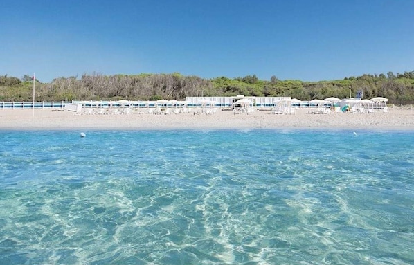 A beachside hotel with clear blue waters, a sandy beach, and a row of white buildings with umbrellas.
