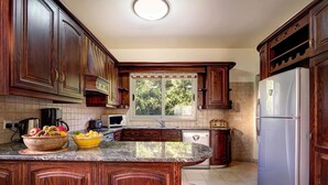 A kitchen with wooden cabinets, a granite countertop, a refrigerator, a dishwasher, and a window with a view of greenery. - Niki Villa (Argaka)