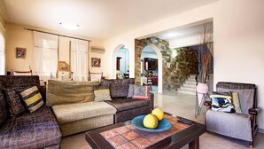 A living room with a sofa, armchairs, a coffee table with fruit, and a view of a kitchen and staircase. - Niki Villa (Argaka)