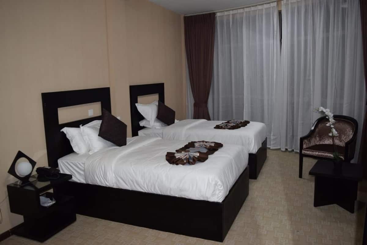 A hotel room with two beds, a nightstand, a chair, and a window with curtains.
