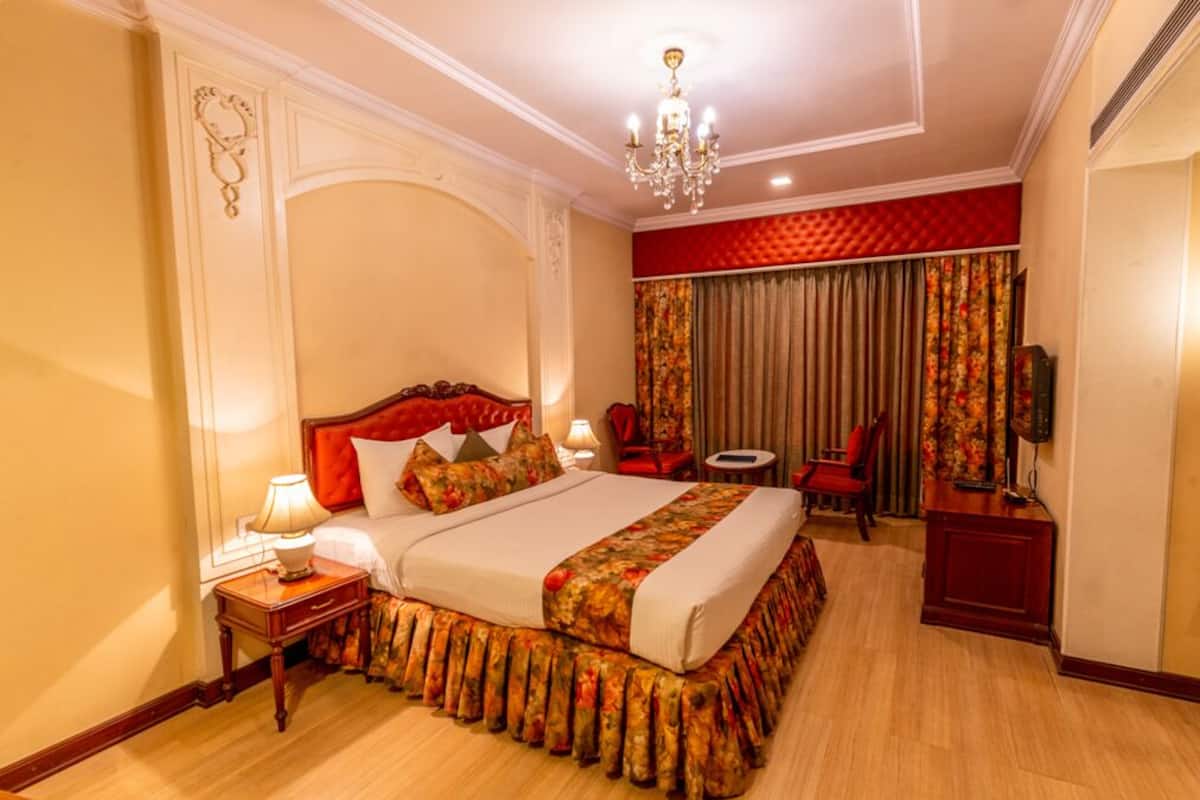 Habitación de hotel con una cama grande, un candelabro, un cabecero tapizado en rojo, una mesita de noche con una lámpara, un sillón rojo, una pequeña mesa con una silla y un televisor sobre un mueble de madera.