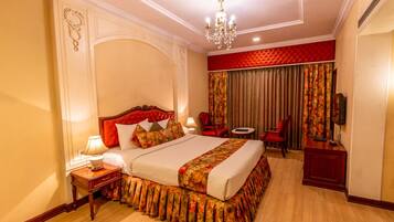 A hotel room with a large bed, a chandelier, a red upholstered headboard, a side table with a lamp, a red armchair, a small table with a chair, and a television on a wooden cabinet.