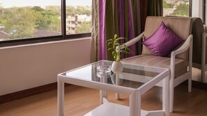 A modern room with a large window, a white chair with a purple cushion, and a glass coffee table. - Hotel Hardeo (Nagpur)