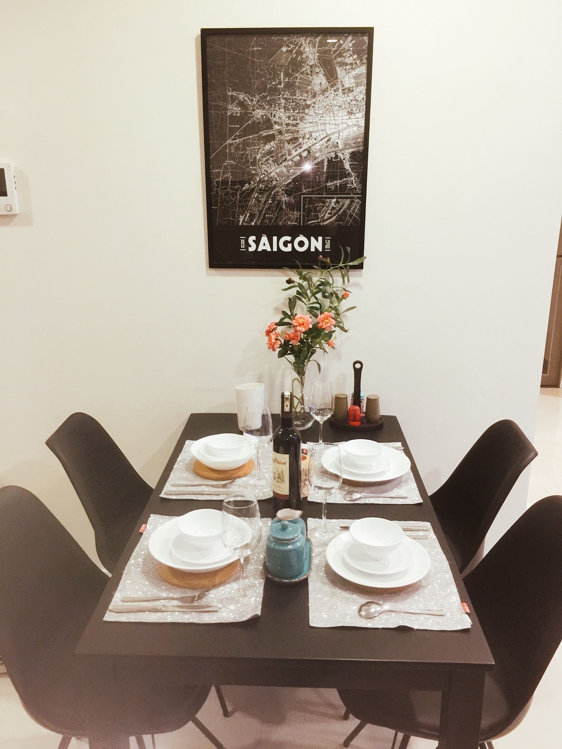 Ett matbord dukat med tallrikar, glas och en vinflaska, med en dekorativ vas och en inramad karta på väggen ovanför.