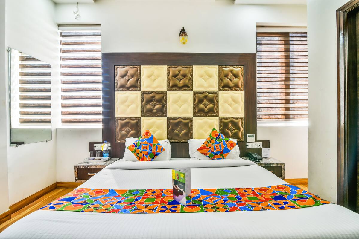 A hotel room with a large bed, a headboard with a geometric pattern, a small bedside table, and a window with blinds.