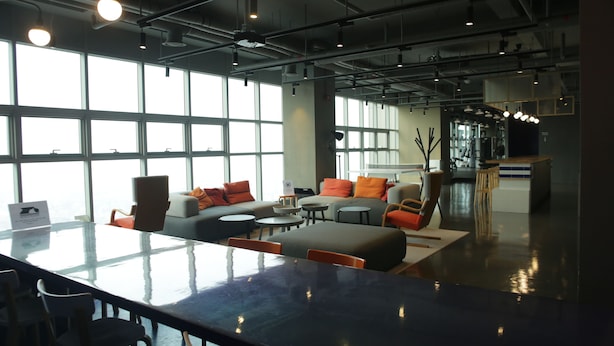 Un espacio de oficina moderno con una gran mesa de cristal, sofás con cojines naranjas y vistas amplias a través de grandes ventanales.