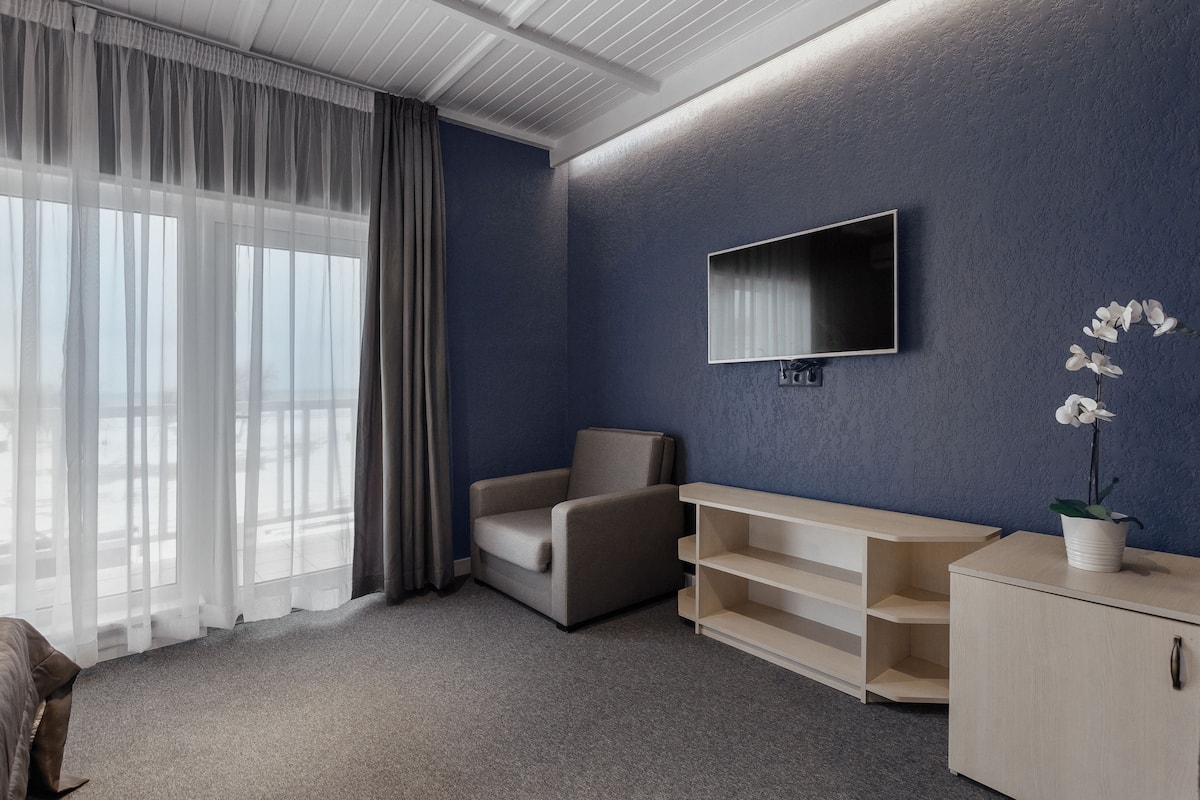 Una habitación de hotel moderna con un ventanal grande, un televisor de pantalla plana montado en la pared, un sillón cómodo, una mesita de noche de madera y una orquídea en maceta.