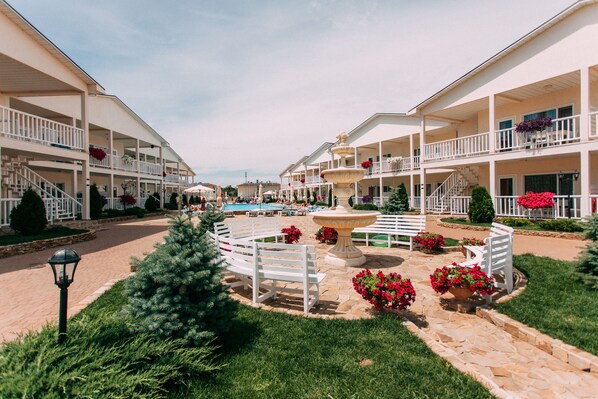 A resort with multiple-story buildings, outdoor swimming pool, and a central fountain surrounded by benches and flower beds.