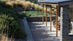 Un espacio al aire libre cubierto con un muro de piedra y vigas de madera, rodeado de vegetación natural y montañas al fondo.