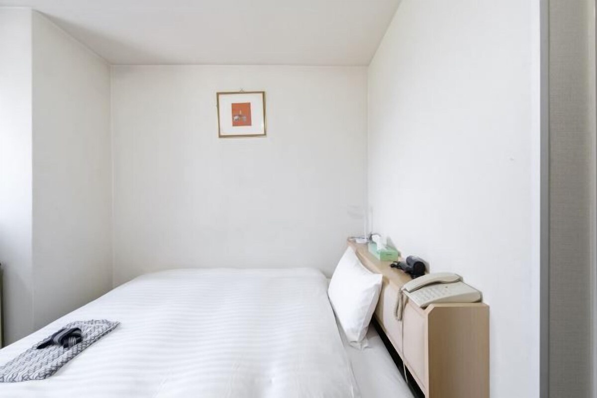 A small, neatly arranged hotel room with a single bed, a bedside table with a lamp, a framed picture on the wall, and a suitcase on the floor.