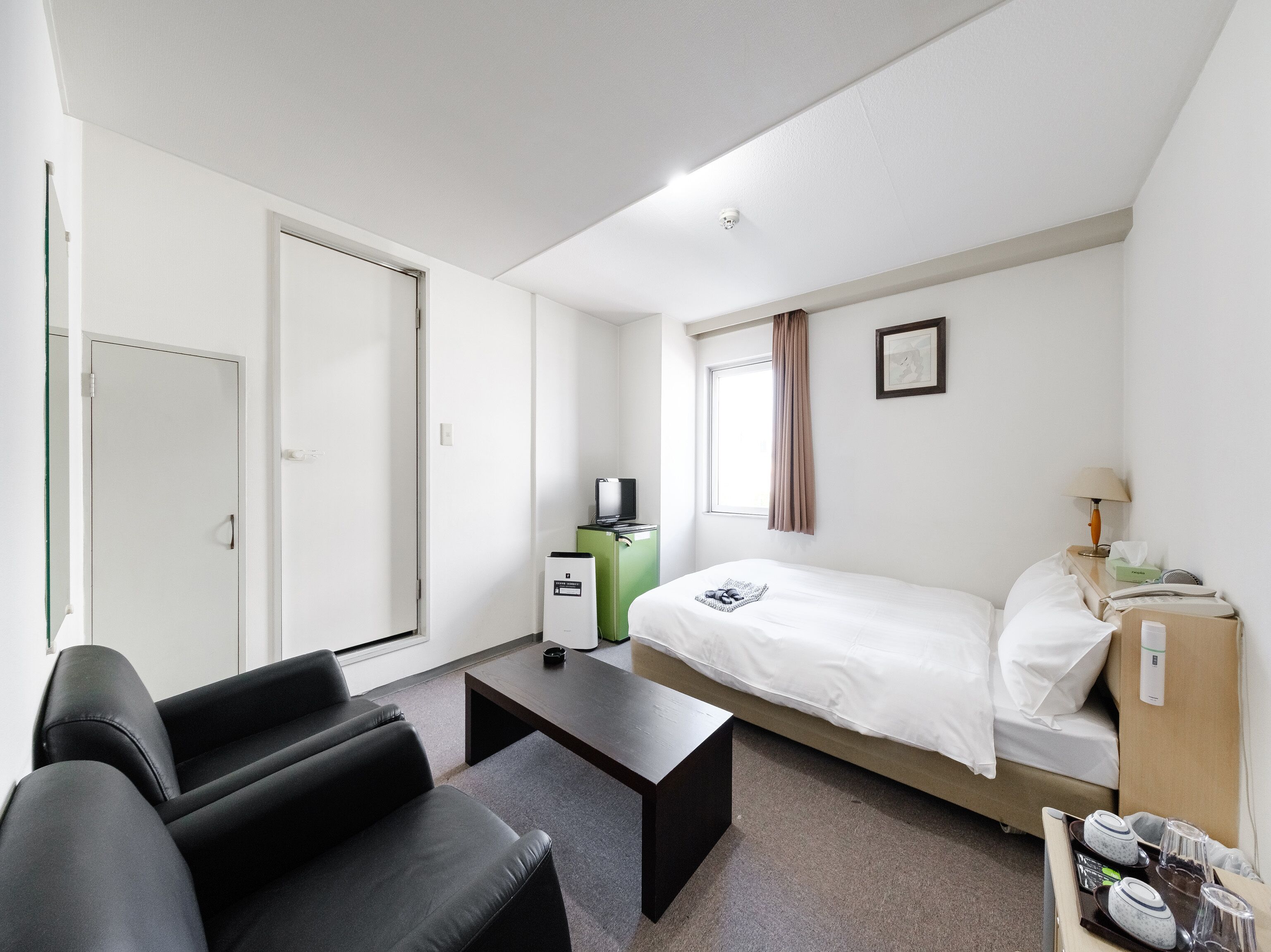 A hotel room with a bed, a desk, a chair, a small table, a nightstand, a lamp, and a window with curtains.
