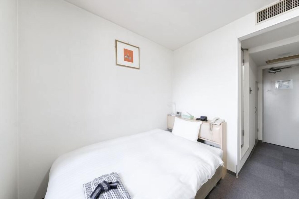 A hotel room with a single bed, a bedside table with a lamp, a small framed picture on the wall, and a door leading to another room.