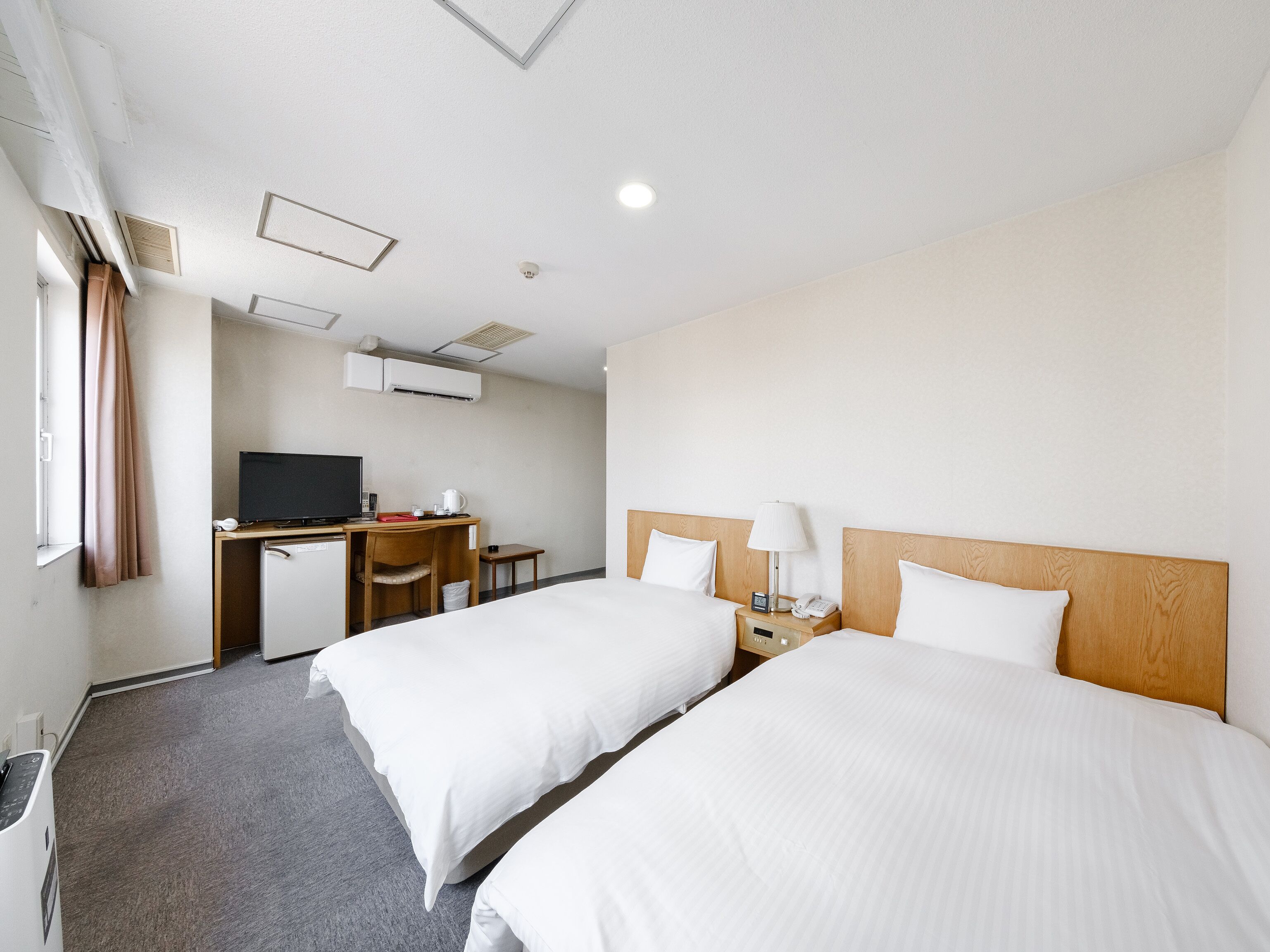 A hotel room with two beds, a desk with a TV, a desk lamp, and a bedside table with a phone and alarm clock.