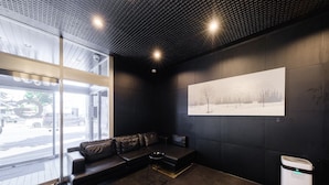 A modern waiting area with a leather sofa, a trash bin, and a large window. - Tabist THE GREEN ASAHIKAWA (Asahikawa)
