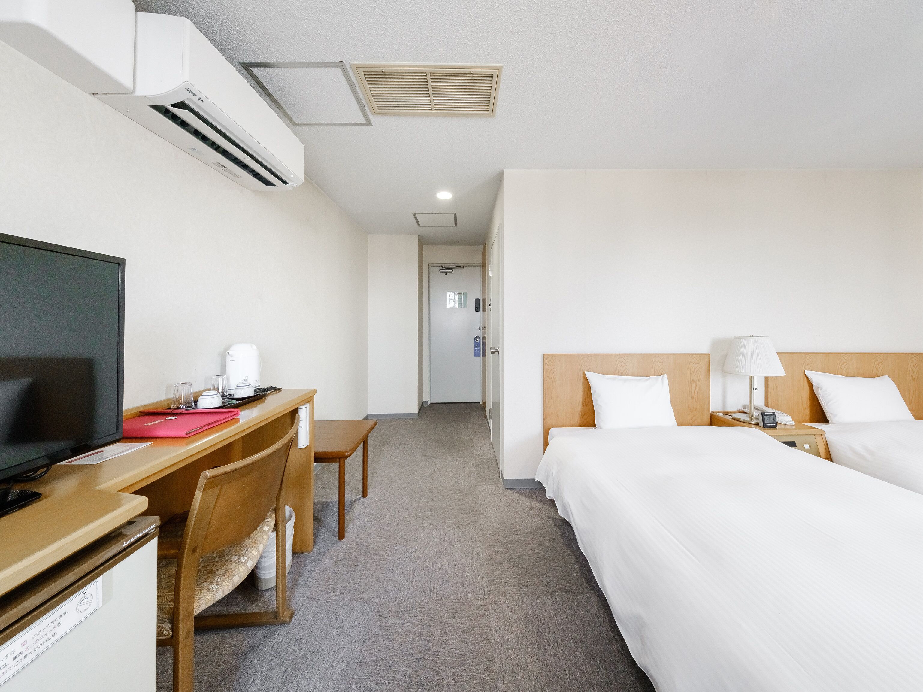 A hotel room with a bed, bedside table, lamp, TV, desk, chair, and an air conditioning unit.