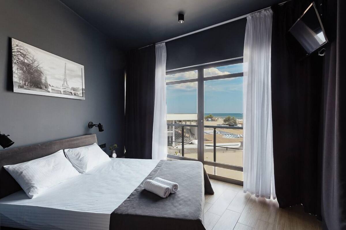 Une chambre d’hôtel avec un grand lit, un tableau représentant la Tour Eiffel, un balcon avec vue sur la plage et un téléviseur à écran plat.