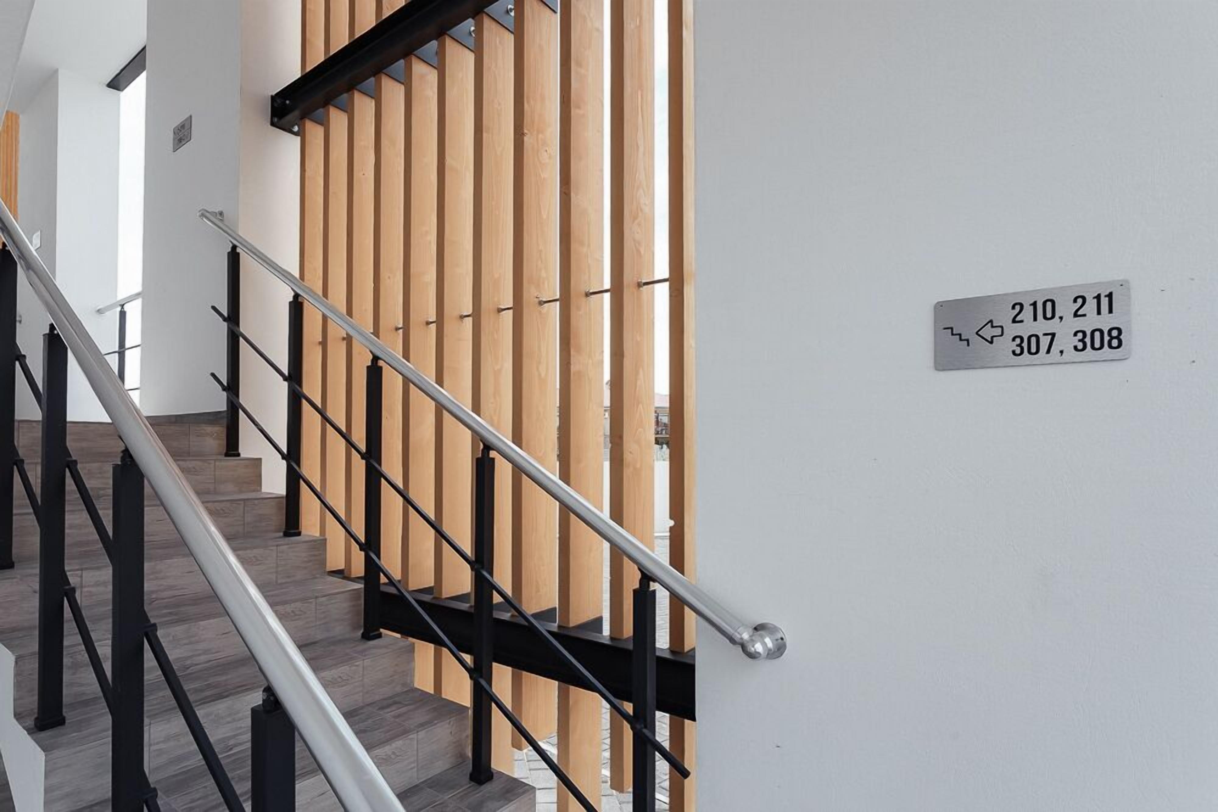 Un escalier avec des rampes noires et des panneaux en bois. Une pancarte indique les numéros des chambres : 210, 211, 307, 308.