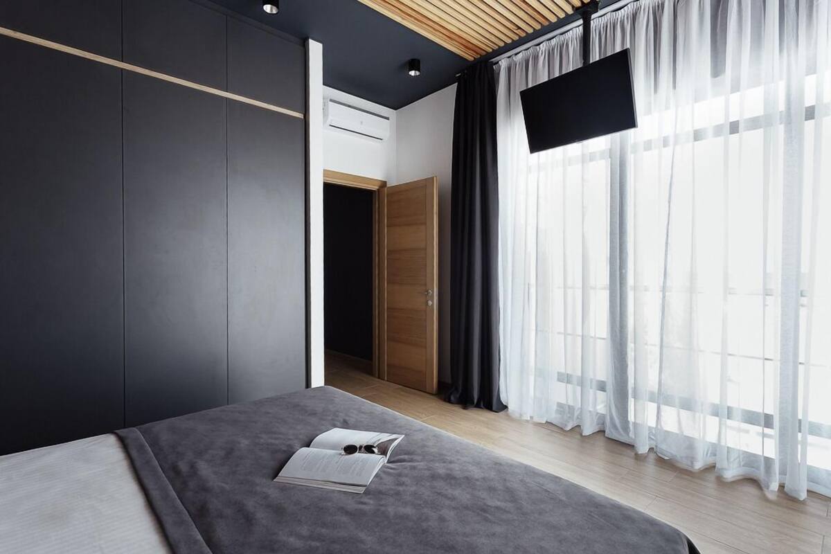 Une chambre moderne avec un lit, une armoire, un téléviseur et une grande fenêtre ornée de rideaux légers.