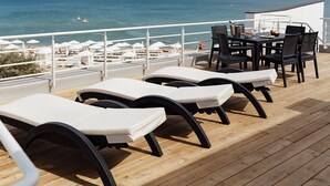 A deck with lounge chairs and a table, overlooking a beach with umbrellas and a view of the sea.
