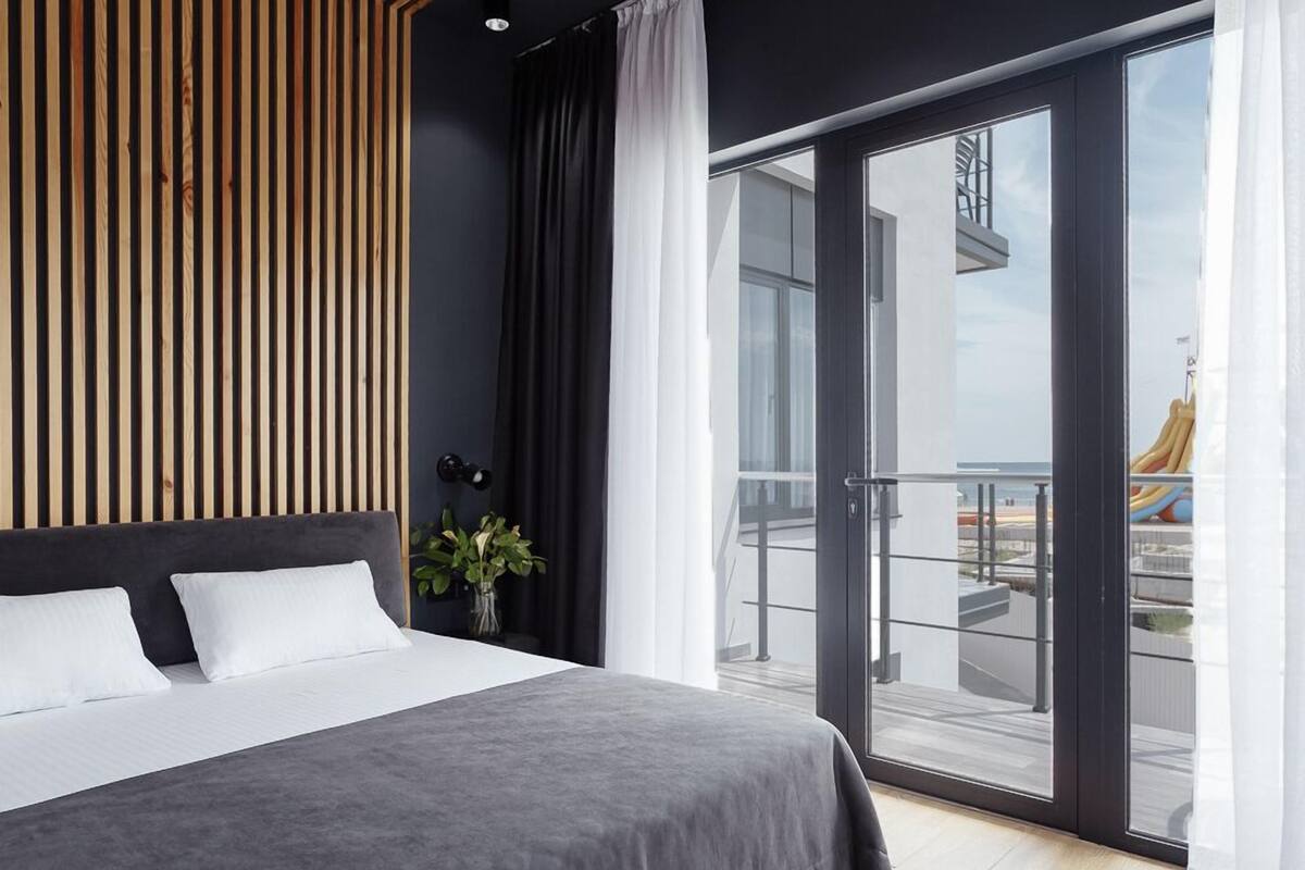 Une chambre moderne avec un grand lit, une tête de lit en lattes de bois, un balcon avec une porte vitrée et une vue sur l’extérieur.